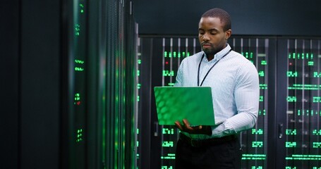 Male system administrator of big data center typing on laptop computer while working in server room. Programming digital operation. Man engineer working online in database center. Telecommunication.