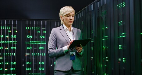 Female system administrator checking big data and tapping on tablet device while working in server...
