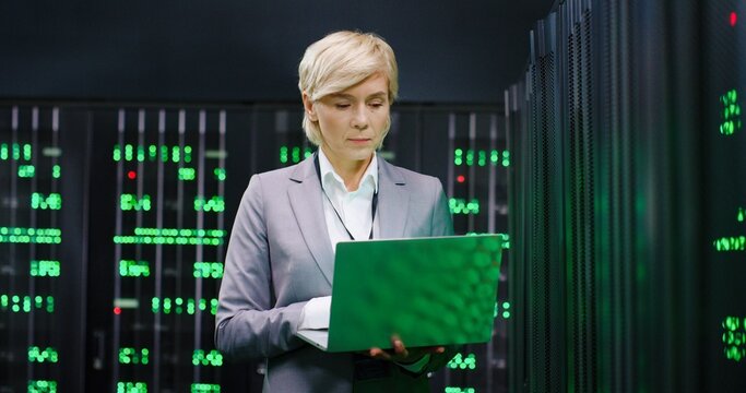 Portrait of beautiful Caucasian female big data specialist wih laptop computer in hands looking at camera in servers room. Professional woman typing on keyboard in database center. Digital storage.