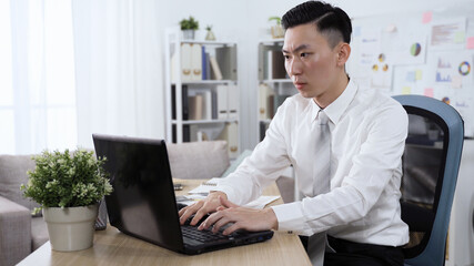 portrait taiwanese male in formal wear is writing a proposal on computer. man pausing and touching his face while thinking hard and proceeding with his paper.