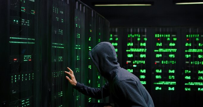 Male Asian guy data thief in dark hood stealing in the server room with tablet device in hands. Digital spy man hacking database. Male hacker commiting cyber crime.