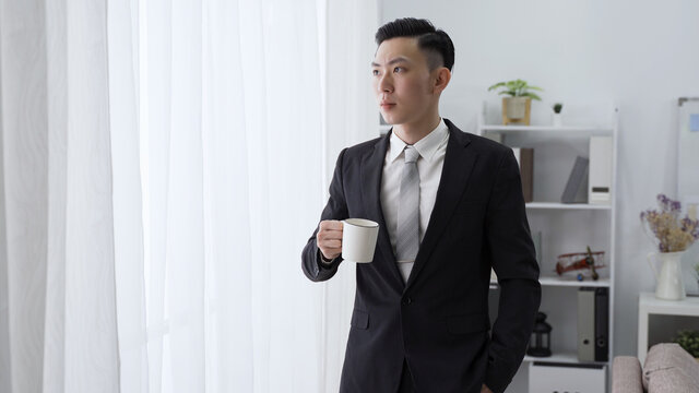male ceo is sipping morning coffee by window with serious expression. asian man in black suit with hand in pocket is looking outside in contemplation