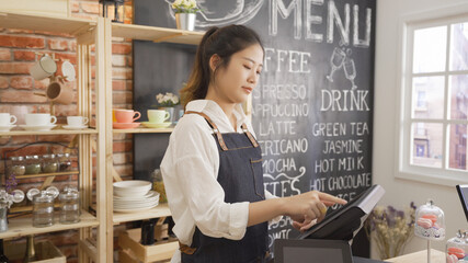 Young girl waitress using tablet computer running her cafe happily smiling waiting for customers. woman barista owner finger touching device screen checking finance of small business coffee shop