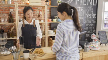 Beautiful waitress serving female customer with cup of coffee take away for breakfast indoors. young barista woman in apron hand showing tasty handmade croissant in counter and recommend to lady