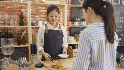 girl baker put croissant on plate to female customer in bread store. young office lady buying breakfast in modern cafe bar. cute waitress in apron working in counter serve client in coffee shop.