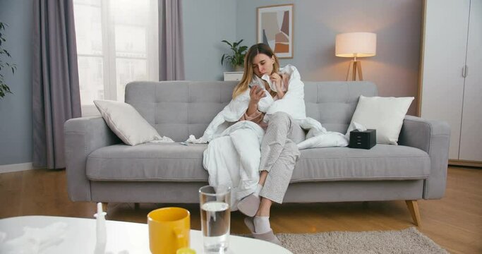 Caucasian sick young woman sitting on couch in living room in blanket, caughing and texting message on smartphone. Ill girl on sofa using mobile phone, tapping and scrolling.