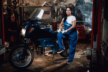 Obraz na płótnie Canvas Beautiful brunette woman in blue overalls posing with big wrench while standing next to a sportbike in garage or workshop