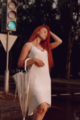 Fototapeta na wymiar a girl in a white dress with red hair with an umbrella walks through puddles after rain at sunset