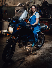Fototapeta na wymiar Beautiful young brunette female mechanic in blue overalls sitting on sportbike in garage or workshop