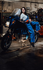Obraz na płótnie Canvas Hot brunette woman in blue overalls posing for a camera while leanign on sportbike in garage or workshop