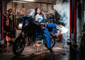 Obraz na płótnie Canvas Hot brunette woman in blue overalls posing for a camera while leanign on sportbike in garage or workshop