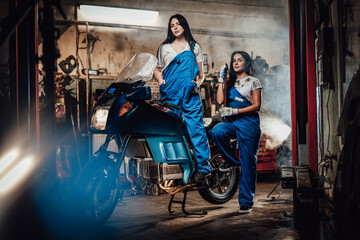 Fototapeta na wymiar Two hot brunette women in blue overalls posing next to a sportbike in authentic workshop garage
