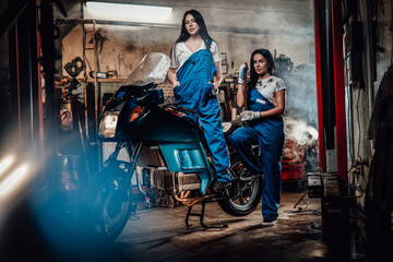Obraz na płótnie Canvas Two hot brunette women in blue overalls posing next to a sportbike in authentic workshop garage