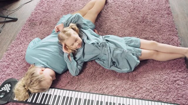 Tired blonde girls relax at home on a pink carpet. Near electronic piano.