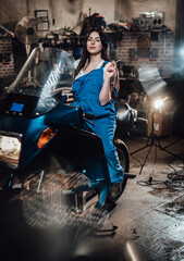 Fototapeta na wymiar Beautiful brunette female mechanic in blue overalls relaxing smoking a cigarette while sitting on sportbike in garage or workshop