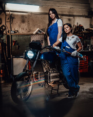 Fototapeta na wymiar Two hot brunette women in blue overalls posing next to a custom bobber in authentic workshop garage
