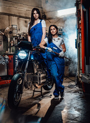 Obraz na płótnie Canvas Two hot brunette women in blue overalls posing next to a custom bobber in authentic workshop garage