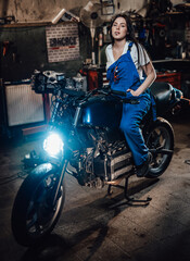 Beautiful brunette female mechanic in blue overalls relaxing smoking a cigarette while sitting on custom bobber in garage or workshop