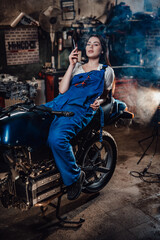 Hot brunette female mechanic in blue overalls relaxing smoking a cigarette while lying on custom bobber in garage or workshop