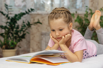 Portrait of a preschool girl who is lying at home on a bed (sofa), reading a book. Education and preparation for school. Free time at home.