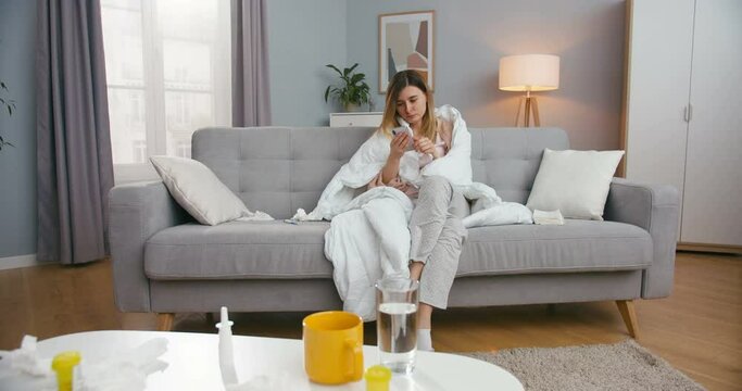 Caucasian unhealthy young woman sitting on couch in living room in blanket with flu, caughing and texting message on smartphone. Ill female on sofa at homeusing mobile phone, tapping and scrolling.