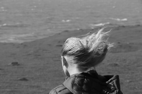 Black and white photo of girl walking away from camera on sandy beach with hair being pulled by the wind 