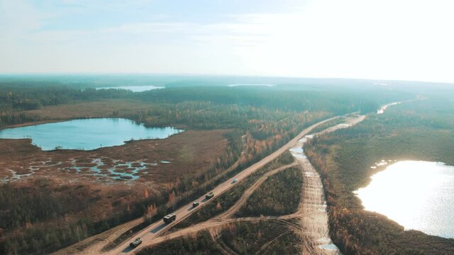A military column of armored vehicles rides through a picturesque landscape among forests and lakes.