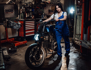 Fototapeta na wymiar Young female mechanic in blue overalls posing with custom bobber in garage or workshop
