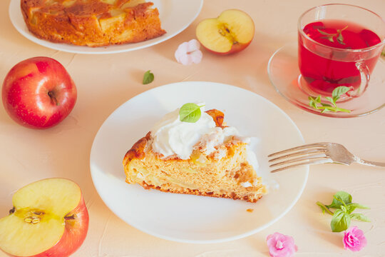 Charlotte apple cooked at home, garnished with whipped cream on a white plate. Around - apples, a cup of tea and tea leaves, flowers, mint leaves. A light image in pastel colors. Home baking concept.