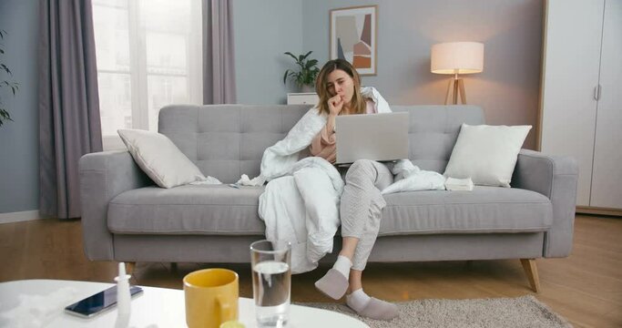 Young Caucasian ill woman under blanket sitting on sofa and typing on laptop computer. Unhealthy girl caughing and having flu or coronavirus while working at home.