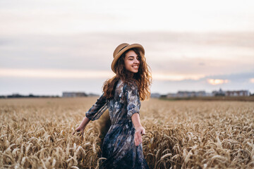Naklejka na ściany i meble Closeup portrait of a beautiful young woman with curly hair. Woman in dress and hat standing in wheat field
