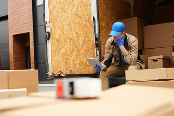 Delivery person in uniform sitting in the car among cardboard boxes and using digital tablet outdoors