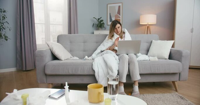 Unhealthy young Caucasian woman with fever sitting on couch in living room and videochatting on laptop. Sick girl caughing and having videochat on computer via webcam. Female talking with video.