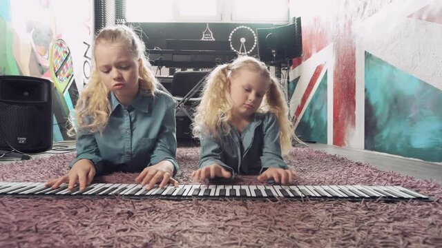 Two cute blonde girls are playing on a synthesizer while lying on the floor.