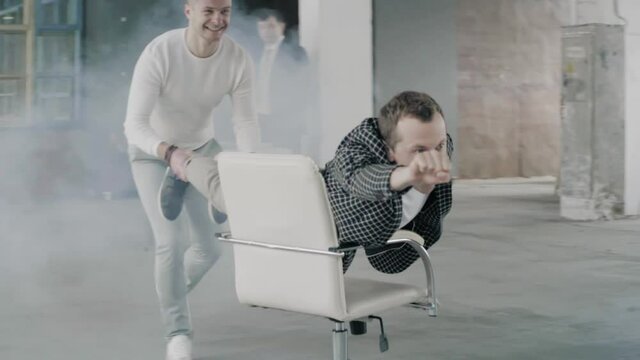 The manager in the office rolls a colleague on a chair like Batman. Office staff having fun.