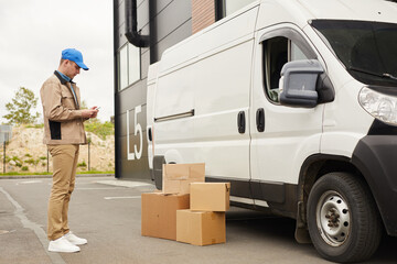 Young man in uniform using his mobile phone while standing near the van and packages outdoors near...