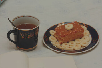 A blue cup of hot tea, a piece of shortbread cake with condensed milk and bananas, cut rings on a saucer and an open book with white blank pages on sheets on the bed.