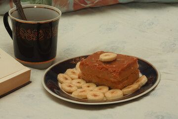 A blue cup of hot tea, a piece of shortbread cake with condensed milk and bananas, cut rings on a saucer and an open book with white blank pages on sheets on the bed.