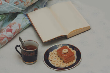 A blue cup of hot tea, a piece of shortbread cake with condensed milk and bananas, cut rings on a saucer and an open book with white blank pages on sheets on the bed.