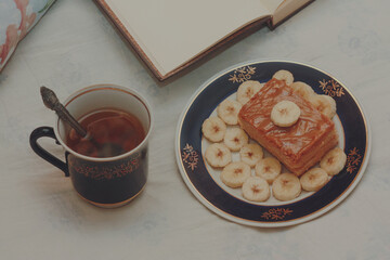 A blue cup of hot tea, a piece of shortbread cake with condensed milk and bananas, cut rings on a saucer and an open book with white blank pages on sheets on the bed.