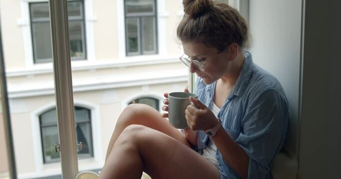 Cosy and casual young woman with hair bun in pajamas sip coffee or tea on balcony or window shelf. Lazy sunday weekend morning in comfortable setting. Me time, relax and unwind, self loving concept