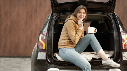 Fototapeta na wymiar Happy brunette with mug sitting in open trunk of black car on summer day