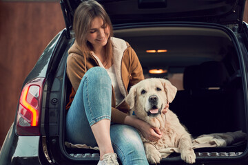 Young woman in blue jeans next to golden retriever sitting in open trunk of black car