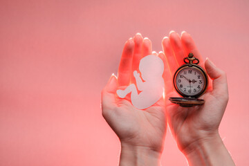 Paper embryo silhouette and clock in woman hands with light. Light coral background with copy...