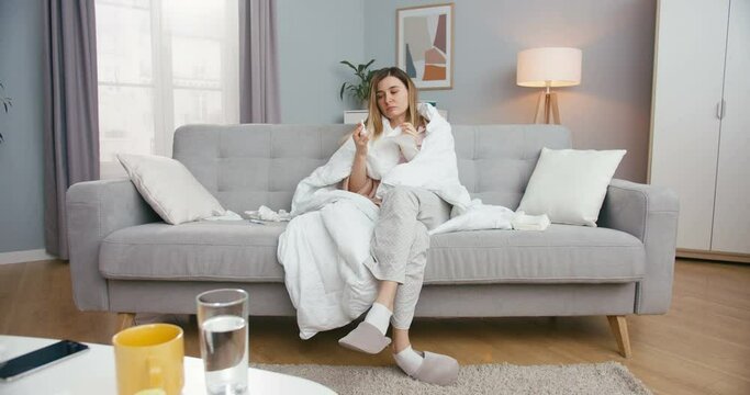 Caucasian young sick female patient spraying medical drops in nose and caughing with napkin while sitting on sofa at home. Ill girl feeling bad and having flu. Covid-19 concept. Unhealthy woman.
