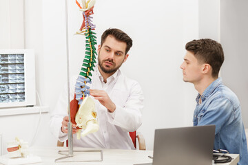 Doctor showing flexible spine model to his patient and explains the cause of his pain in medical...