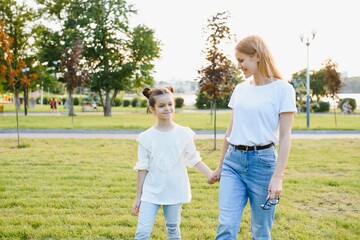 Attractive young woman with her little cute daughter are spending time together outdoors. Mom with daughter in park on a green grass during the sunset.