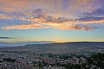 Dawn in the valley of Cappadocia. The sun rises from behind the mountains on the horizon. The sky is blue, the clouds are painted in pink, gold, lilac tones. In the valley is a village .