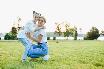 Fototapeta premium Attractive young woman with her little cute daughter are spending time together outdoors. Mom with daughter in park on a green grass during the sunset.