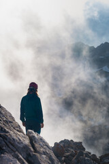 Persona de espaldas asomada al precipicio desde una cumbre con luces de la niebla y montañas de fondo